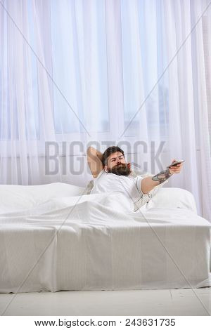 Man In Shirt Laying On Bed, Watching Tv, White Curtains On Background. Macho With Beard Holds Contro