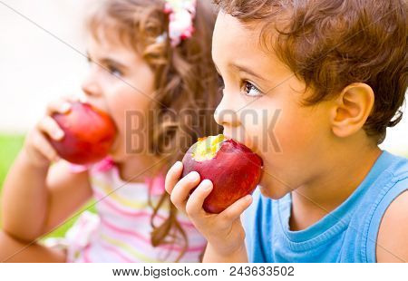 Photo of two happy children eating apples, brother and sister having picnic outdoors, cheerful kids biting red ripe peach, adorable infant holding fresh fruits in hands, healthy nutrition concept 