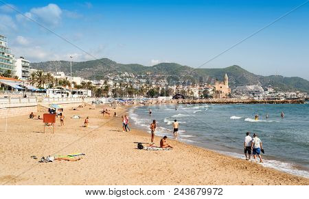 SITGES, SPAIN - OCTOBER 12, 2017: People relaxing, sunbathing or bathing in the Mediterranean sea, at the main beach of Sitges, highlighting the Sant Bartomeu i Santa Tecla Church in the background