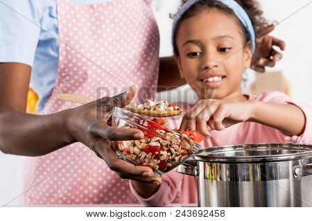 African American Mother And Daughter Putting Ingredientes In Saucepan On Kitchen