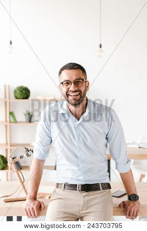 Happy portrait of businesslike man wearing white shirt sitting on table in office and smiling at camera