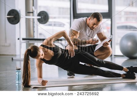 Male Personal Trainer Looking At Timer And Young Athletic Woman Doing Side Plank On Fitness Mat