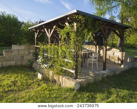 Brown Tiber Wooden Gazebo Or Pergola With Climbing Plants And Flowers, Table And Chairs And Surround