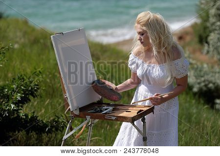 Beautiful Girl Draws A Picture In The Park Using A Palette With Paints And A Spatula. Easel And Canv