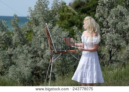 Beautiful Girl Draws A Picture In The Park Using A Palette With Paints And A Spatula. Easel And Canv