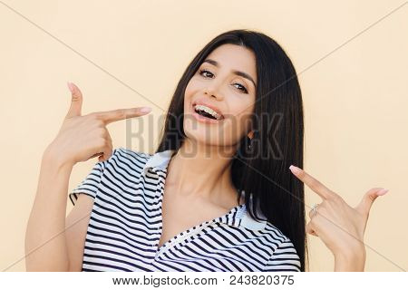 Portrait Of Happy Gorgeous Female Model Indicates With Fore Fingers At Her Face, Advertises Braces F