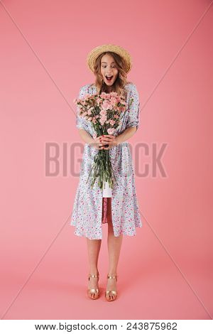 Full length portrait of a cheerful young woman in summer dress and straw hat posing while holding carnations bouquet isolated over pink background