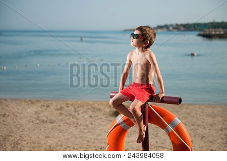 Cute Small Kid In Sunglasses Sitting On Beach With Lifebuoy Outdoor Summer Like Baywatch Movie Heroe