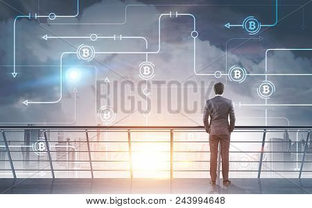 Young Businessman Standing On A Skyscraper Balcony Looking At Bitcoin Network Hologram In A Morning 