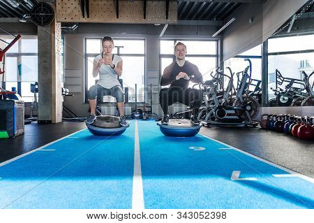 Fit Couple Working On Bosu Ball In Fitness Studio. Doing A Squat Exercise