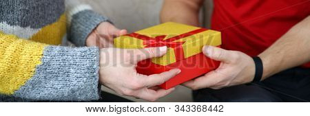 Close-up View Of Female Presenting Gift To Another Person. Nice Red Box With Yellow Top And Bow. Man