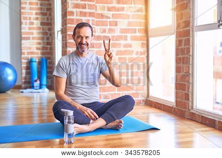 Middle age handsome sportman sitting on mat doing stretching yoga exercise at gym showing and pointing up with fingers number two while smiling confident and happy.