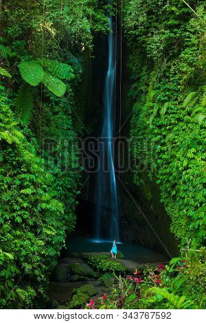 Young Traveler Woman At Waterfall In Tropical Forest. Caucasian Woman Wearing Blue Dress. View From 