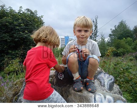 A Boy And A Girl Are Brother And Sister. Children With Blond Hair Walk Next To Trees And Bushes. Gir