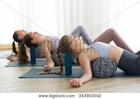 Restorative Yoga With A Bolster. Group Of Three Young Sporty Attractive Women In Yoga Studio, Lying 