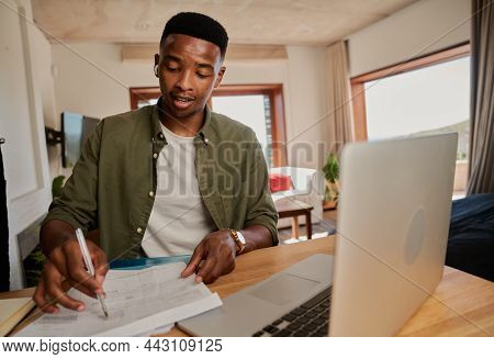 Young Adult Black Male Wearing Earphones Making Notes While On Online Meeting. Sitting At Kitchen Co