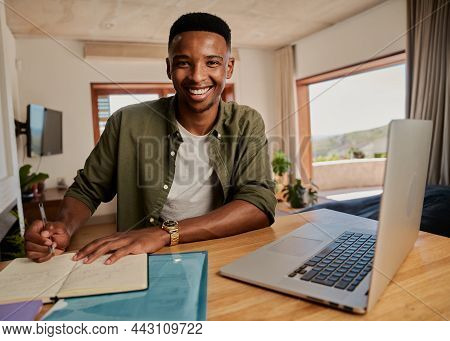 Portrait Of Young Adult Black Male Writing Note In Notebook Next To Laptop. Smiling At Camera, Sitti