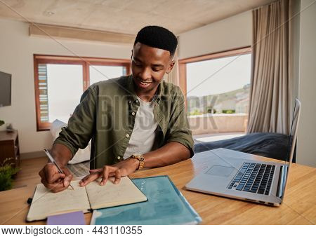 Young Adult Black Male Writing Note In Notebook Next To Laptop, Smiling. Sitting In Modern Apartment
