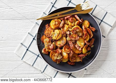 Bratkartoffeln, Fried Potatoes With Bacon And Onion On A Plate With Cutlery On A White Wooden Table,