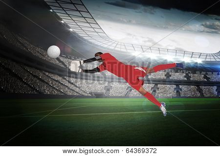Goalkeeper in red making a save in a large football stadium with lights