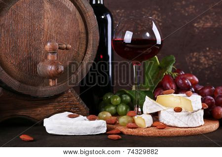 Supper consisting of Camembert cheese, wine and grapes on cutting board and wine barrel on wooden table on brown background