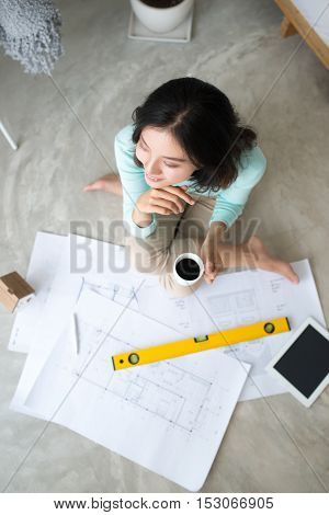 Work everywhere. Confident young beautiful asian woman working with blueprint while sitting on the floor at home