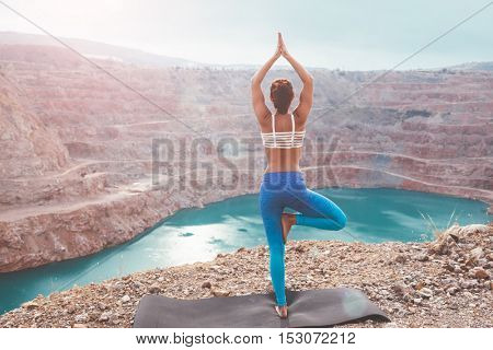 Young sporty girl wearing blue activewear stretching outdoors. Woman training yoga pose on mountain hill, calm and silent view.