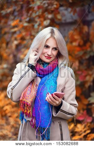 Young beautiful girl listening to MP3 player in autumn park. Beautiful autumn woman listening to music in autumn park enjoy autumn day.Beautiful young blond girl with headphones and smart phone in park in autumn. Cool young woman listening to music outdoo