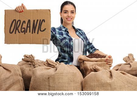 Cheerful female farmer posing with a pile of burlap sacks filled with coffee beans and a cardboard sign that says organic isolated on white background