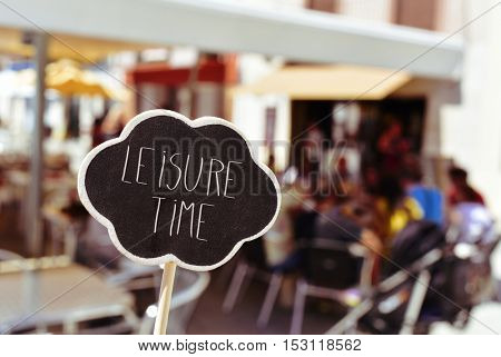 closeup of a thought bubble-shaped signboard with the text leisure time written in it, in front of the blurred terrace of a restaurant