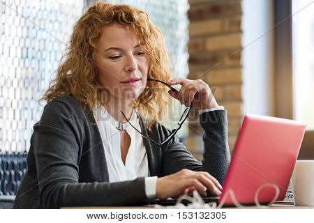 Absorbing work. Pretty ginger lady sitting at table in cafe, holding glasses at mouth and looking at her modern laptop.
