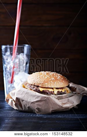 burger on a paper package served with glass full of ice waiting for a drink. At a dark wooden background. Close view. Vertical shot