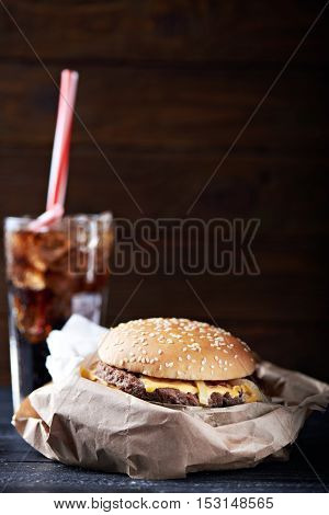 burger on a paper package served with a glass full of ice and drink. At a dark wooden background. Close view. Vertical shot