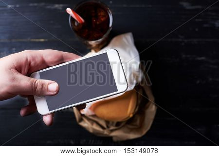 Man using smartphone with empty screen with blur cheeseburger on a background. Dark photo