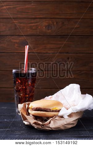 Small graceless burger on a paper package served with a glass full of ice and drink. At a dark wooden background. Close view. Vertical shot
