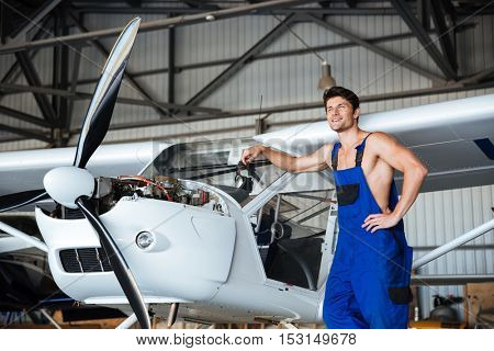 Happy handsome mechanic in overall resting in front of the plane in a shed