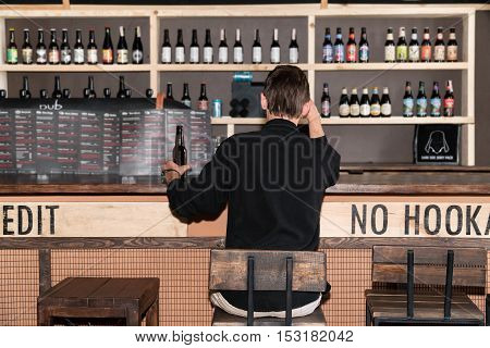 Lonely man in black shirt sitting with his back to the camera at the bar holding a bottle of beer