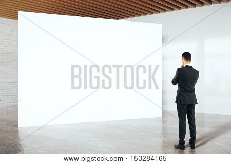 Thoughtful businessman in suit looking at blank billboard in white birck interior. Mock up 3D Rendering