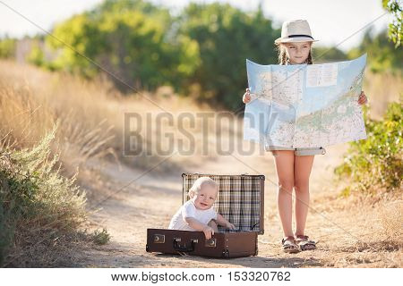 Little boy in a white t-shirt is resting sitting in a large open brown suitcase on a country road, the elder sister is studying map of the area, a girl 6 years old in a gray summer suit with long braids, in a light hat and a beige backpack