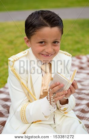 Young boy with white sailor suit in his First Communion smiling with a prayer book and rosary in his hands.