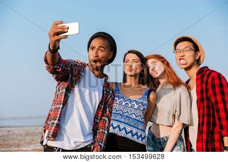 Group of amusing young friends taking selfie with cell phone and making funny faces outdoors