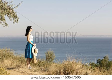 Young woman (brunette) in a blue skirt and hat looks at sea. Selective focus.