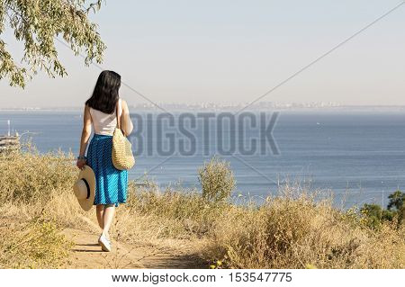 Young woman (brunette) in a blue skirt and hat looks at sea. Selective focus.