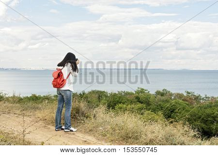 Young girl (brunette) in jeans with a red backpack with a camera taking pictures of the sea. Selective focus.