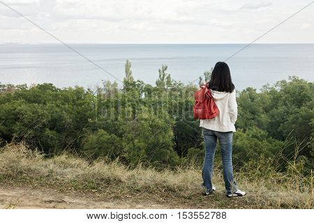 Young girl (brunette) and jeans with a red backpack looking out to the sea. Selective focus.