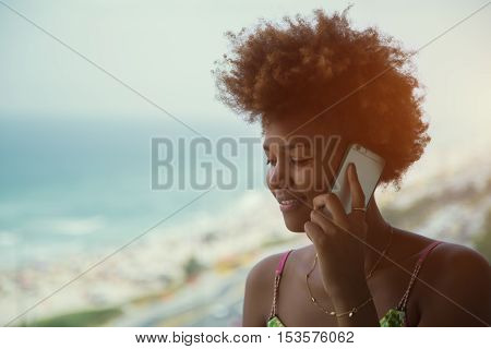 Young black pretty happy attractive female with curly hair speaking on a mobile phone standing near tropical ocean sunny bright summer day vintage colour filter