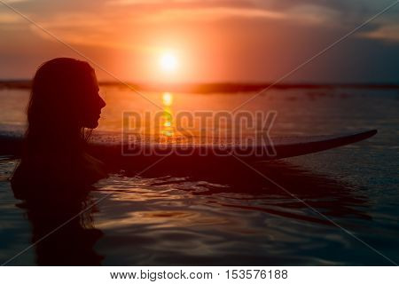 Silhouette of surfer girl standing almost fully in water in bikini with surfboard on tropical beach at beautiful sunset
