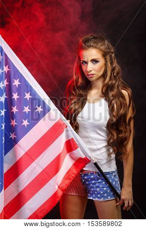 Young attractive girl holding a US flag on the flagpole. She a T-shirt and shorts with a print of the flag. Long curly hair. Portrait of a real patriot. Stars and Stripes.