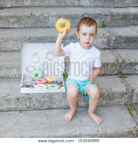 kid holding a donut. boy sitting on the steps with a box of colorful glazed donuts. little child playing with a donut. concept of junk food for kids