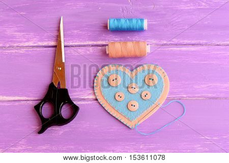 How to make a felt heart crafts. Step. Join the felt edges of felt heart with blue thread. Scissors, thread, needle on wooden background. Crafts idea for Valentine's day, wedding, mother's day